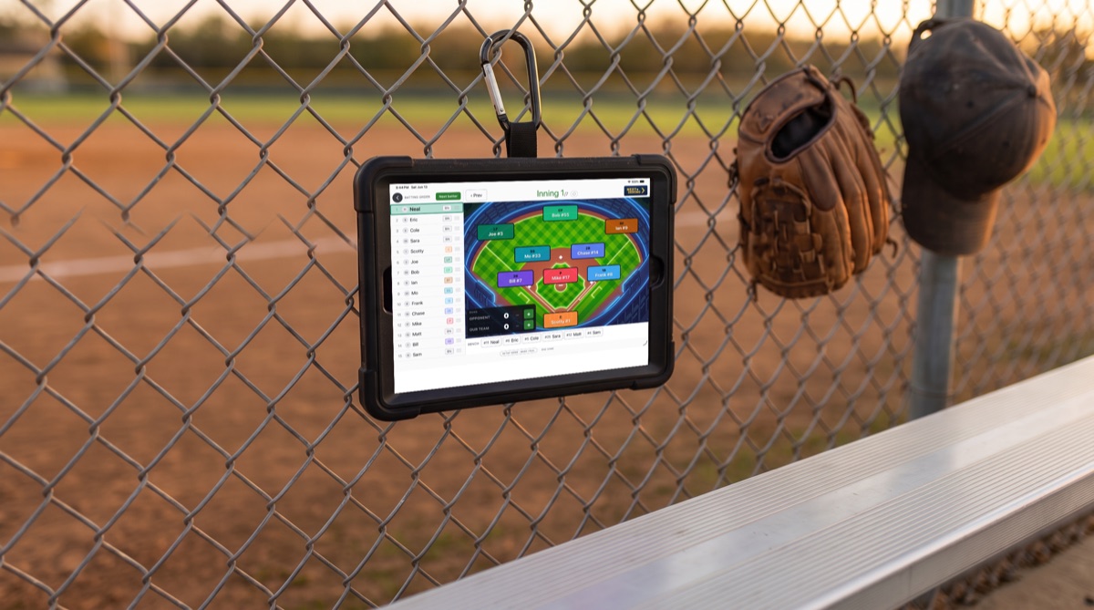 A tablet running Next Inning clipped to a chain-link dugout fence at a youth baseball field, showing the batting order beside the fielding diamond